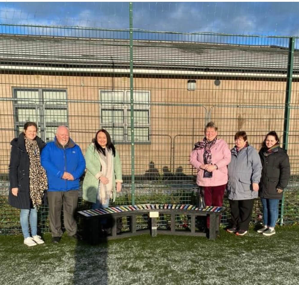 Group of people of all ages that are a part of Maynooth Disability Access Group standing at a bench .